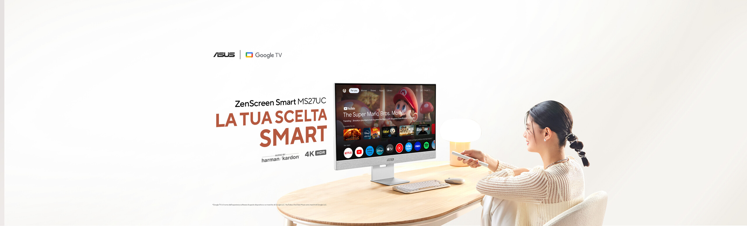 L'immagine mostra una persona seduta di fronte a un monitor ASUS, modello ZenScreen Smart MS27UC, su un tavolo di legno chiaro. La persona sembra utilizzare un telecomando per interagire con il monitor, che mostra un'interfaccia di Google TV con varie app e contenuti video. Sullo schermo sono visibili icone di diverse applicazioni.