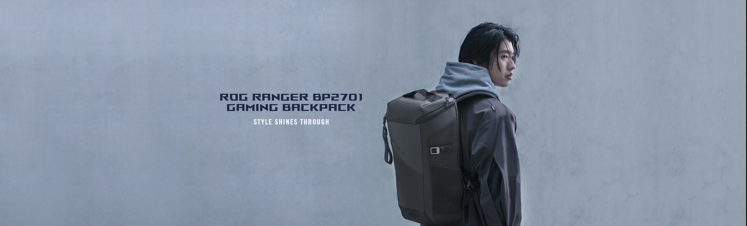 A person with a dark jacket standing near a concrete wall, looking over his shoulder and wearing an ROG Ranger BP2701 Gaming Backpack.